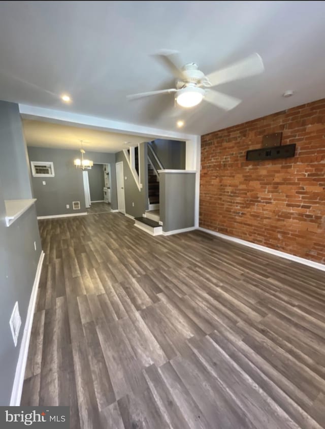 unfurnished living room with brick wall, dark wood-type flooring, and ceiling fan with notable chandelier