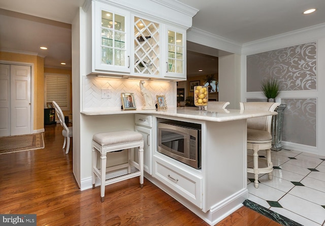 bar featuring stainless steel microwave, decorative backsplash, crown molding, white cabinetry, and light hardwood / wood-style flooring