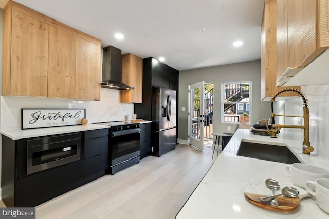 kitchen with wall chimney range hood, appliances with stainless steel finishes, light stone countertops, light hardwood / wood-style flooring, and sink