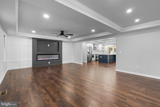 unfurnished living room with a raised ceiling, crown molding, ceiling fan, and dark wood-style flooring