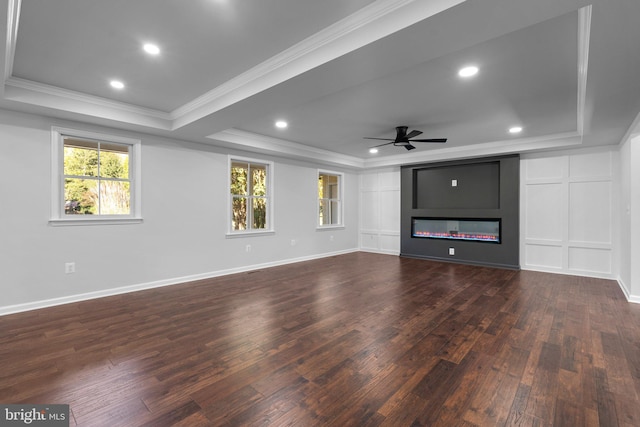 unfurnished living room featuring a ceiling fan, a tray ceiling, dark wood-type flooring, a glass covered fireplace, and ornamental molding