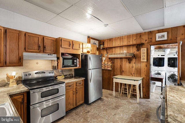 kitchen with light stone counters, stainless steel appliances, stacked washer and dryer, and wood walls