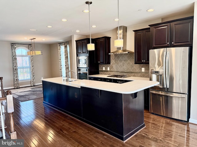 kitchen featuring dark wood-type flooring, hanging light fixtures, wall chimney exhaust hood, an island with sink, and stainless steel appliances