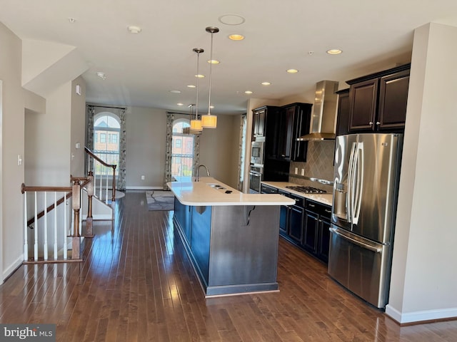 kitchen with a kitchen bar, appliances with stainless steel finishes, a kitchen island with sink, wall chimney range hood, and hanging light fixtures