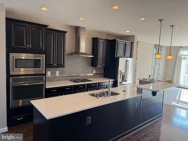 kitchen with appliances with stainless steel finishes, dark hardwood / wood-style flooring, wall chimney exhaust hood, a kitchen island with sink, and hanging light fixtures