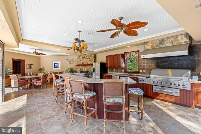 kitchen with wall chimney range hood, a stone fireplace, a kitchen breakfast bar, appliances with stainless steel finishes, and ceiling fan