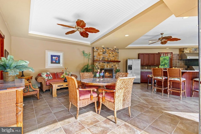 dining area with ceiling fan, a raised ceiling, ornamental molding, and a stone fireplace