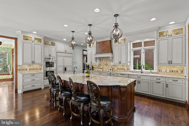 kitchen with stainless steel double oven, a kitchen island with sink, dark hardwood / wood-style floors, hanging light fixtures, and custom exhaust hood