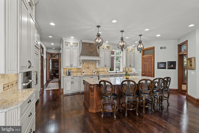 kitchen featuring custom exhaust hood, hanging light fixtures, light stone countertops, a kitchen island with sink, and dark wood-type flooring