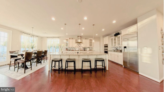 kitchen with wall chimney exhaust hood, decorative light fixtures, built in fridge, and a large island with sink