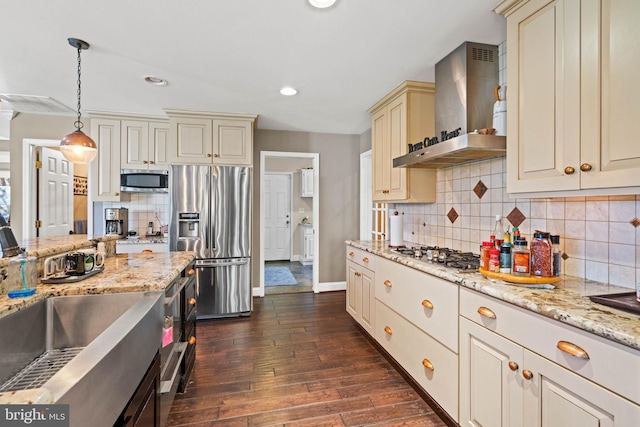 kitchen featuring dark wood-type flooring, cream cabinets, hanging light fixtures, wall chimney exhaust hood, and stainless steel appliances