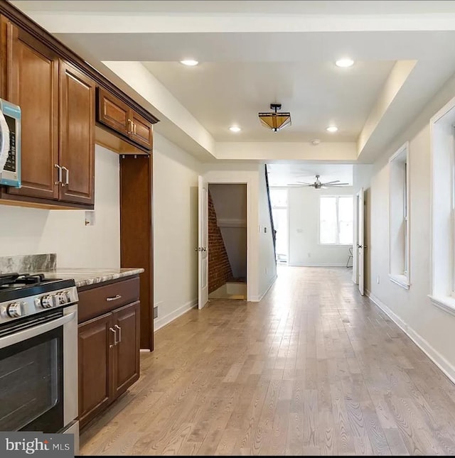 kitchen featuring light stone countertops, light wood-type flooring, a raised ceiling, stainless steel range with gas cooktop, and ceiling fan