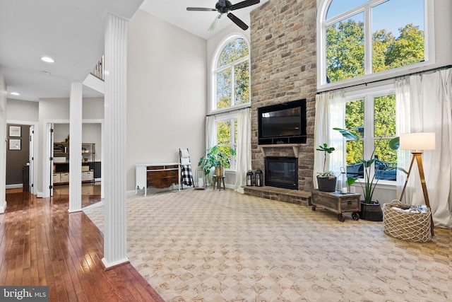 living room with a towering ceiling, wood-type flooring, a stone fireplace, ornate columns, and ceiling fan