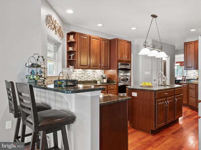 kitchen with dark wood-type flooring, a kitchen island with sink, hanging light fixtures, and dark stone counters