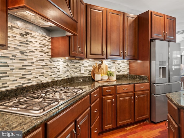 kitchen with stainless steel appliances, backsplash, dark stone countertops, hardwood / wood-style floors, and custom exhaust hood