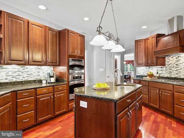 kitchen with sink, an island with sink, pendant lighting, dark wood-type flooring, and decorative backsplash