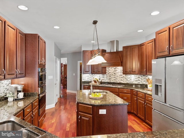 kitchen featuring custom exhaust hood, appliances with stainless steel finishes, a kitchen island with sink, hardwood / wood-style flooring, and pendant lighting