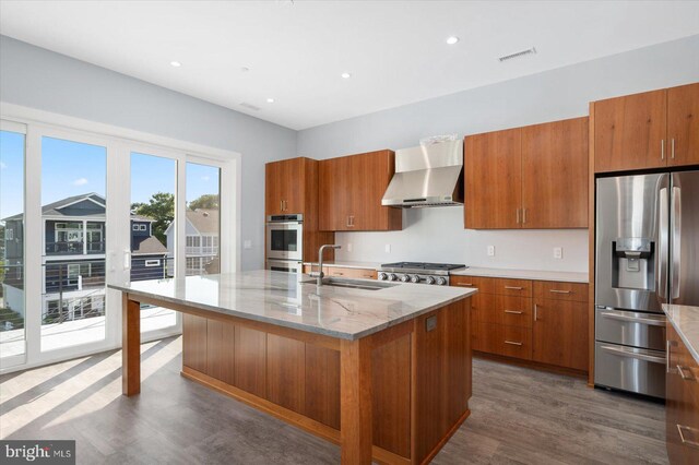 kitchen with wall chimney range hood, appliances with stainless steel finishes, a kitchen island with sink, dark hardwood / wood-style floors, and sink