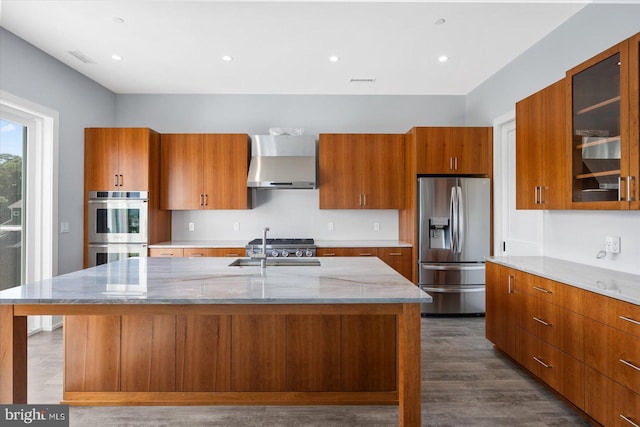 kitchen with a spacious island, wall chimney range hood, appliances with stainless steel finishes, light stone counters, and dark hardwood / wood-style flooring