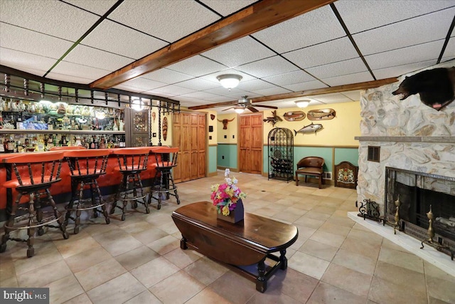 bar featuring ceiling fan, a stone fireplace, a drop ceiling, and tile patterned flooring