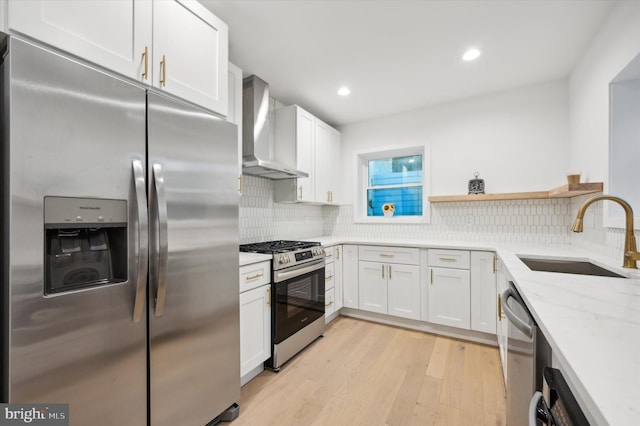 kitchen with wall chimney range hood, appliances with stainless steel finishes, white cabinetry, light hardwood / wood-style floors, and sink