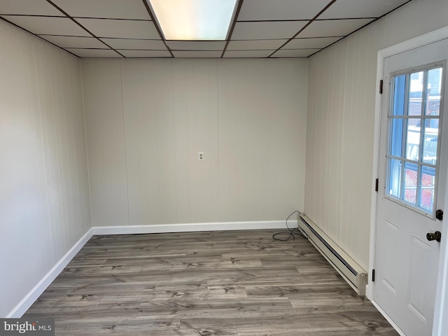 empty room featuring a drop ceiling, wood-type flooring, baseboard heating, and a healthy amount of sunlight