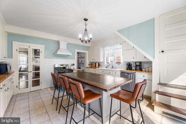 kitchen with wall chimney range hood, appliances with stainless steel finishes, butcher block countertops, white cabinetry, and decorative light fixtures