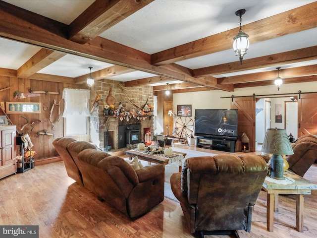 living room with a stone fireplace, wood walls, a barn door, beamed ceiling, and light hardwood / wood-style flooring