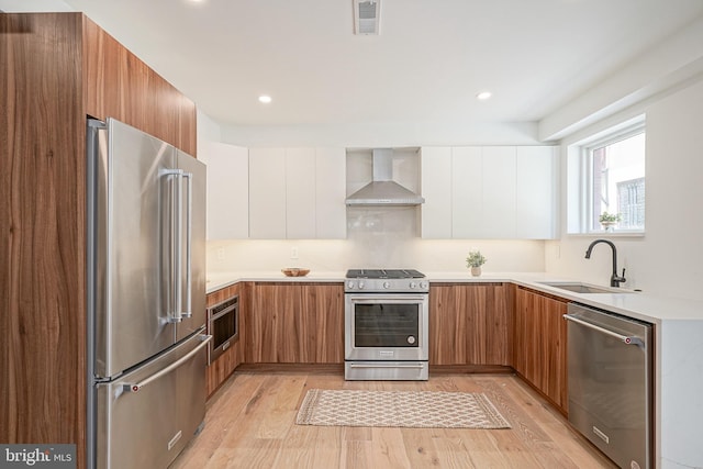 kitchen with appliances with stainless steel finishes, wall chimney range hood, sink, white cabinetry, and light hardwood / wood-style flooring