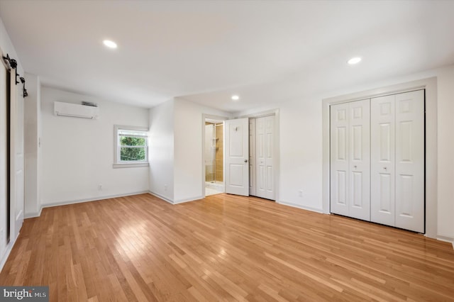 unfurnished bedroom featuring an AC wall unit, light hardwood / wood-style flooring, a barn door, connected bathroom, and multiple closets