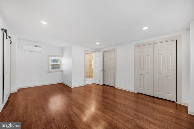 unfurnished bedroom featuring a wall mounted air conditioner, ensuite bathroom, two closets, wood-type flooring, and a barn door