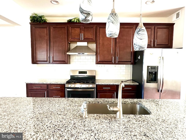 kitchen with wall chimney range hood, appliances with stainless steel finishes, light stone countertops, sink, and decorative light fixtures