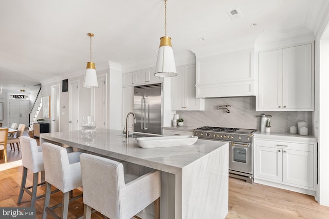 kitchen with light stone counters, premium appliances, a center island with sink, white cabinets, and hanging light fixtures