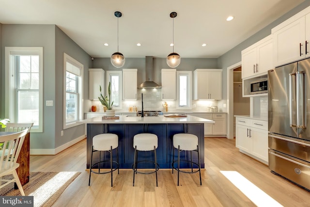 kitchen with pendant lighting, wall chimney range hood, stainless steel appliances, an island with sink, and white cabinets