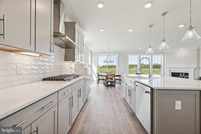 kitchen featuring appliances with stainless steel finishes, wall chimney exhaust hood, decorative light fixtures, gray cabinets, and a center island with sink