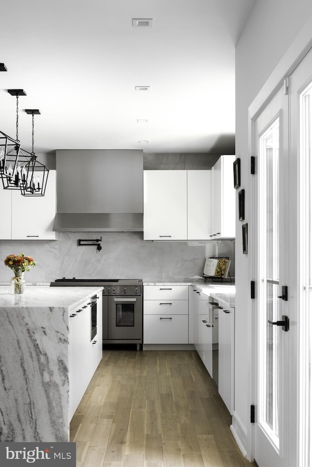 kitchen featuring light wood-type flooring, white cabinetry, wall chimney exhaust hood, pendant lighting, and a notable chandelier