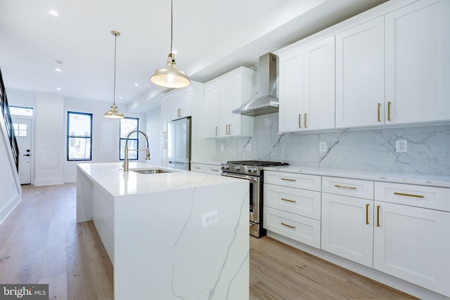 kitchen featuring a center island with sink, sink, wall chimney range hood, appliances with stainless steel finishes, and decorative light fixtures