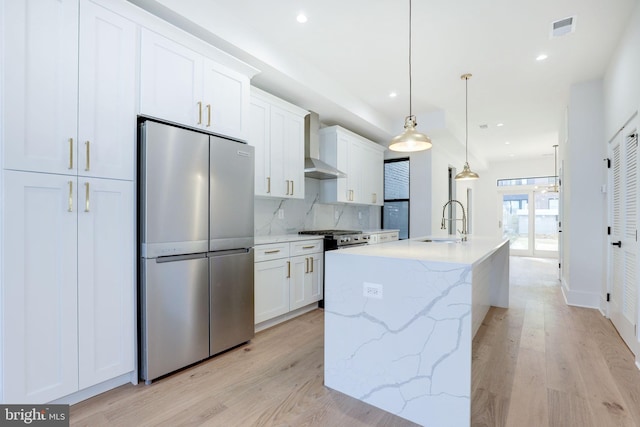 kitchen featuring sink, an island with sink, wall chimney range hood, white cabinetry, and appliances with stainless steel finishes