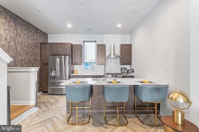 kitchen with wall chimney range hood, a kitchen bar, kitchen peninsula, stainless steel fridge, and light parquet floors