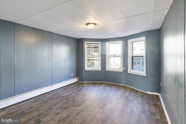 empty room with baseboard heating, wood walls, a drop ceiling, and dark hardwood / wood-style flooring