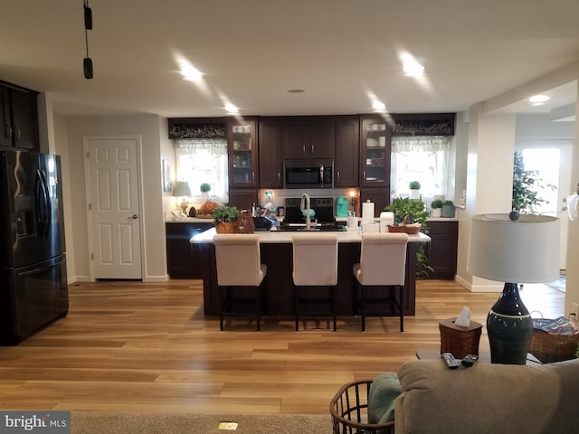 kitchen with dark brown cabinetry, an island with sink, a kitchen bar, black appliances, and light wood-type flooring