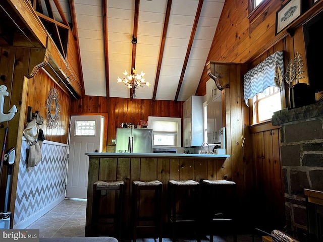 kitchen with vaulted ceiling with beams, kitchen peninsula, stainless steel fridge, a notable chandelier, and wood walls