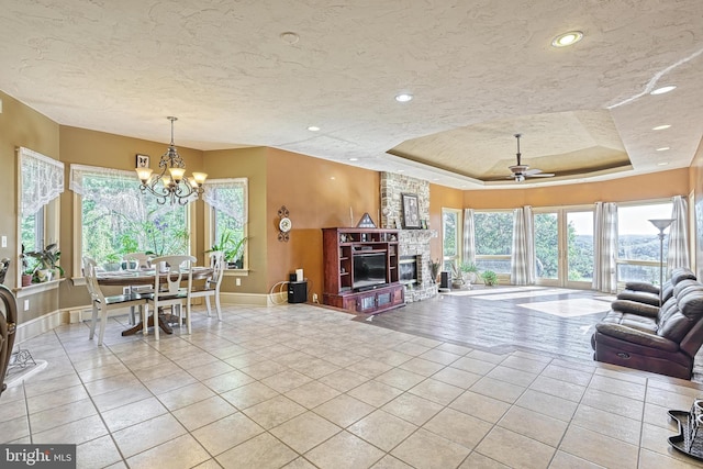 tiled living room featuring a fireplace, a textured ceiling, a tray ceiling, and ceiling fan with notable chandelier