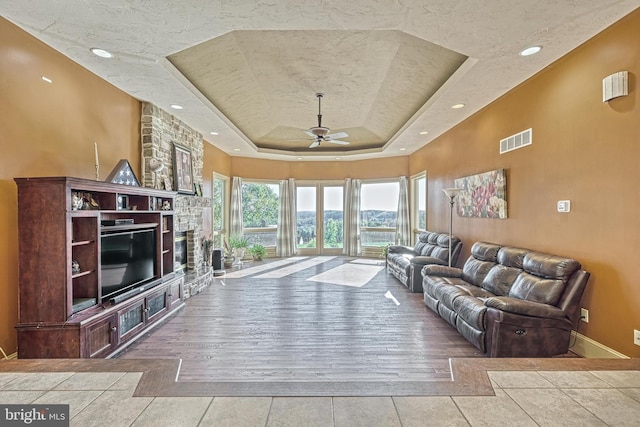 living room with ceiling fan, a raised ceiling, a textured ceiling, light wood-type flooring, and a stone fireplace