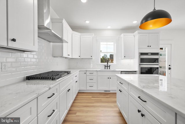 kitchen featuring wall chimney range hood, hanging light fixtures, stainless steel appliances, white cabinets, and light hardwood / wood-style flooring