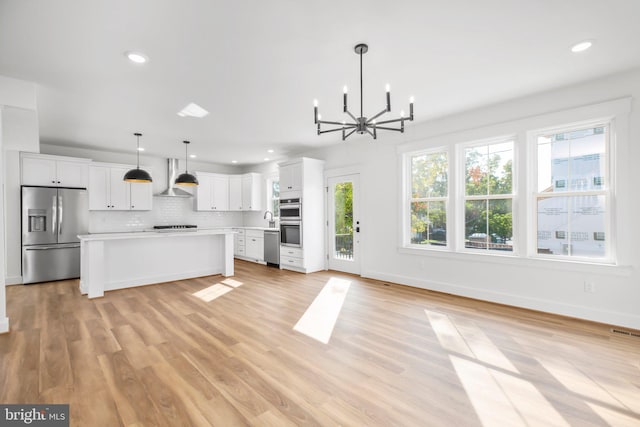 kitchen featuring wall chimney range hood, hanging light fixtures, stainless steel appliances, white cabinetry, and light hardwood / wood-style floors