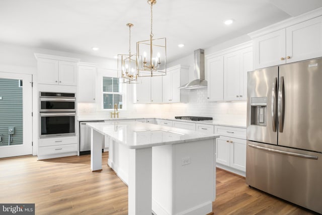 kitchen with appliances with stainless steel finishes, a kitchen island, wall chimney exhaust hood, pendant lighting, and white cabinets