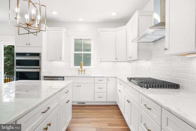 kitchen with light wood-type flooring, stainless steel appliances, wall chimney exhaust hood, decorative light fixtures, and white cabinets