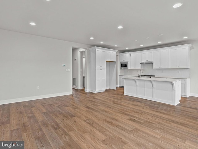 kitchen featuring stainless steel microwave, white cabinets, decorative backsplash, a kitchen island with sink, and light wood-type flooring