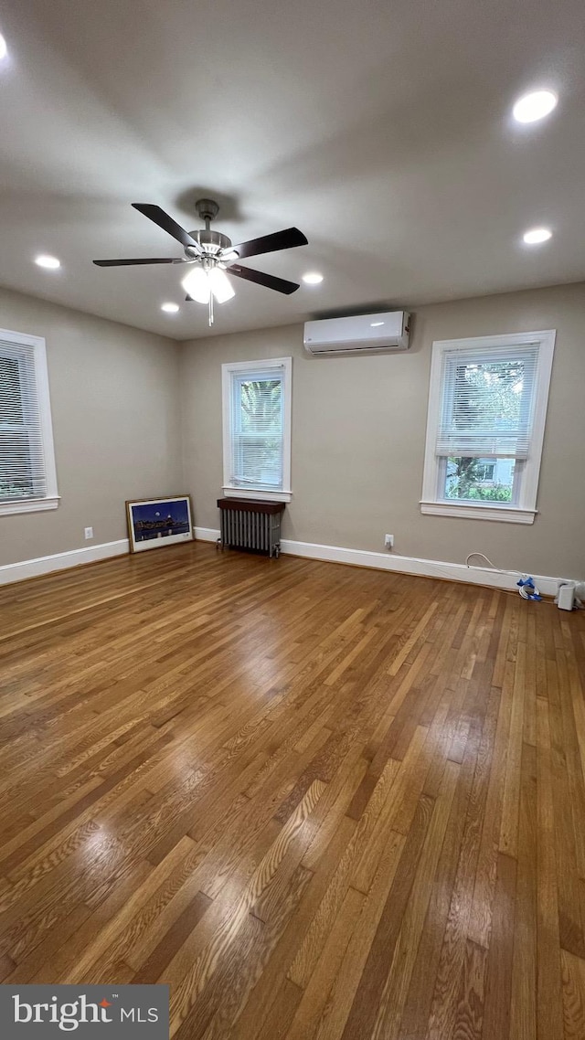 unfurnished living room featuring an AC wall unit, hardwood / wood-style floors, radiator heating unit, and ceiling fan